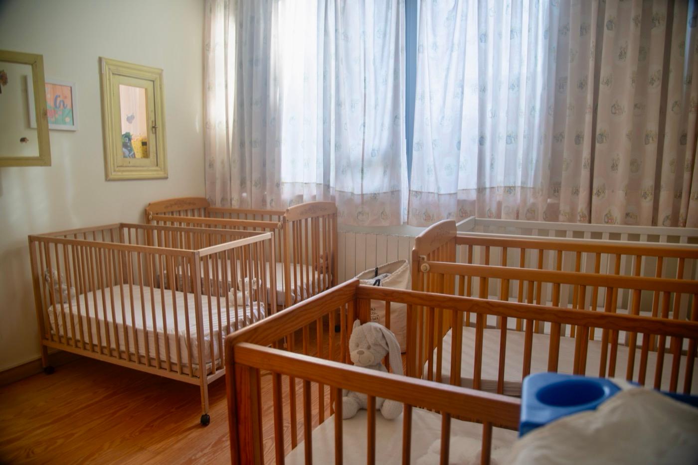 Aula de bebés: las cunas de madera junto a los ventanales con cortinas